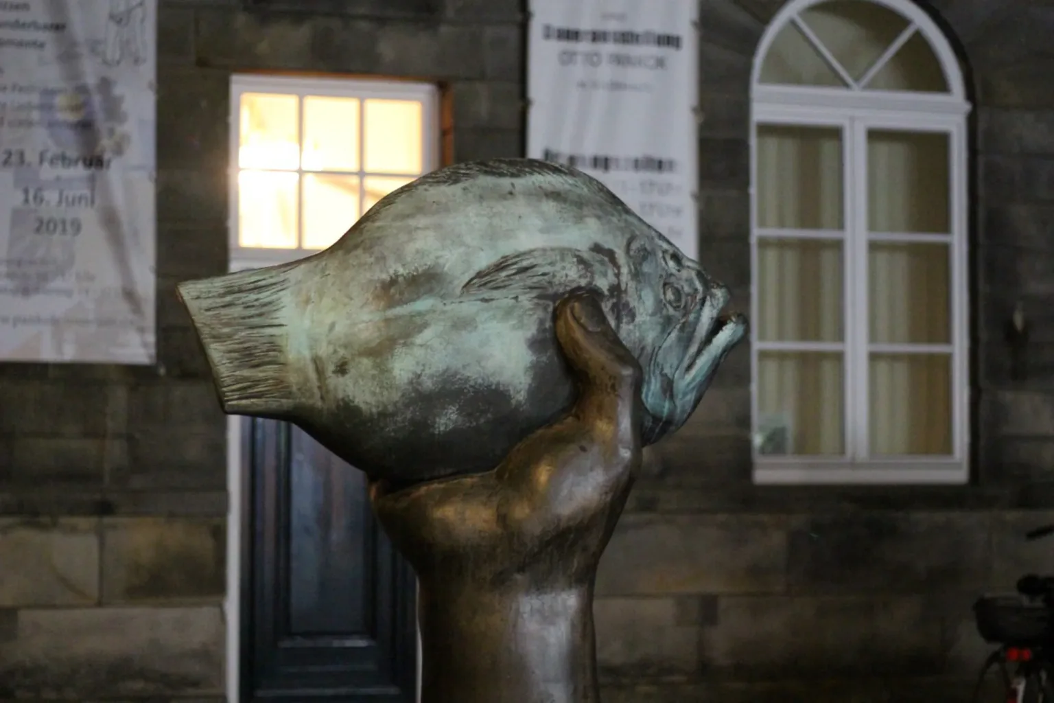 Eine Statue eines Fisches, der von einer menschlichen Figur gehalten wird, wahrscheinlich die Statue einer Person. Die Fischstatue ist recht groß und nimmt einen großen Teil des Bildes ein. Die Statue befindet sich vor dem Museum.