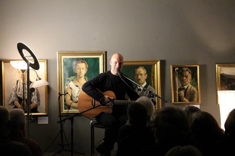 Ein Mann sitzt auf einem Hocker in einem Raum und spielt Gitarre. Er ist von einer Gruppe von Menschen umgeben, die ihn beim Spielen beobachten. In der Szene sind mindestens zehn Personen zu sehen, von denen einige näher an dem Mann stehen und andere weiter weg. Das Publikum scheint engagiert zu sein und die Darbietung zu genießen. Der Raum enthält auch mehrere Gemälde, die zur Atmosphäre und zum Ambiente der Szenerie beitragen.